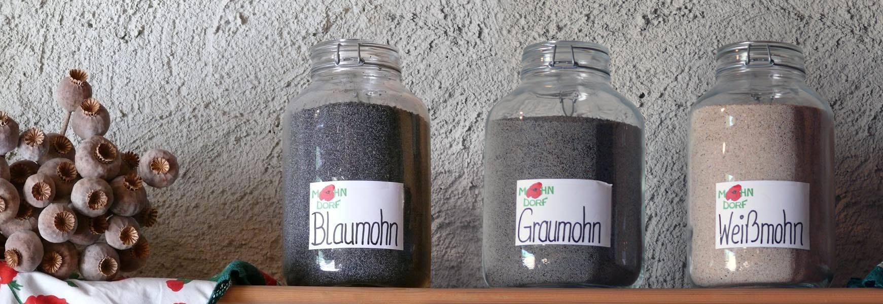 Tres frascos con semilla de amapola azul, gris y blanca, junto a cápsulas secas de amapola