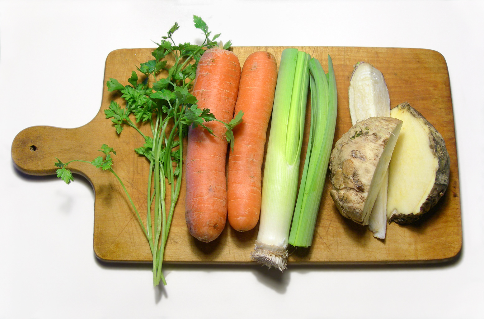 Verduras para caldo sobre una tabla de madera: perejil, dos zanahorias, un poro y un apio nabo partido