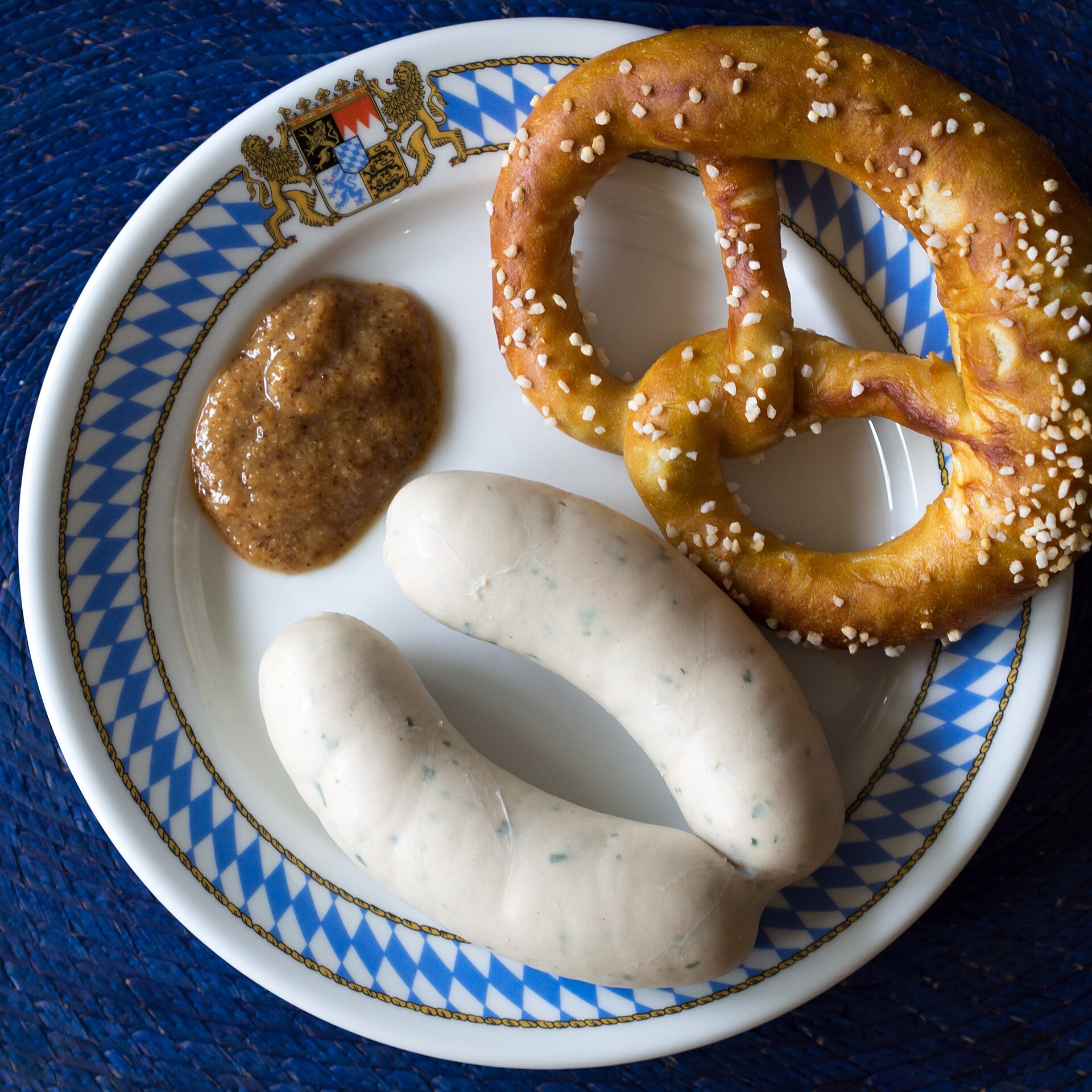 Dos Weißwürste con brezel y una cucharada de mostaza dulce gruesa en plato azul y blanco